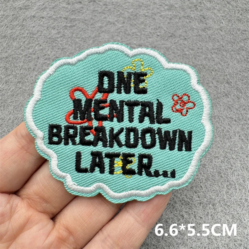 A teal embroidered patch with black text reading ONE MENTAL BREAKDOWN LATER and a gold crown detail, held in a hand.