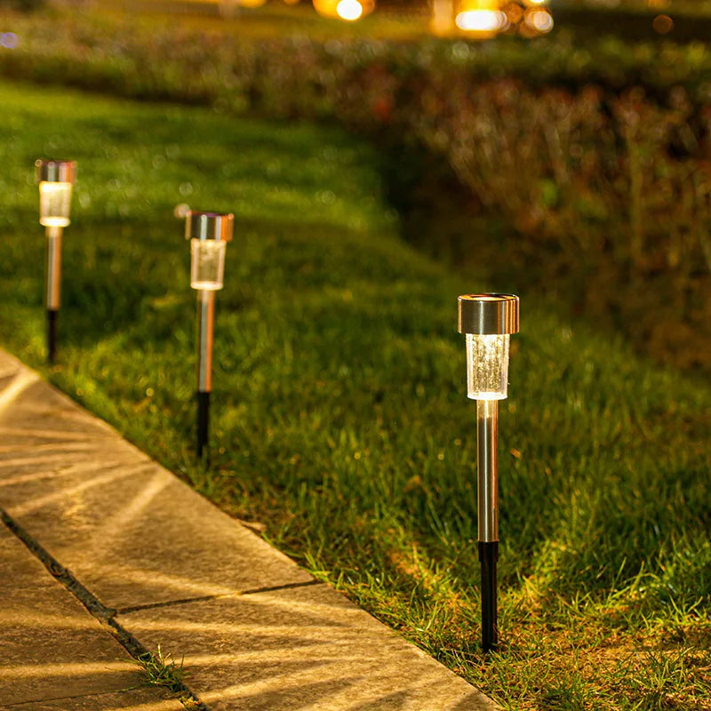 A row of modern solar powered garden lantern lights standing in a grassy area next to a paved walkway.