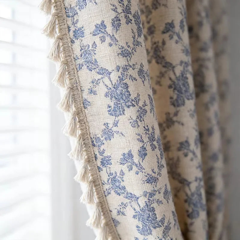A close-up of a cream and blue floral cotton-linen curtain fabric with fringed edges.