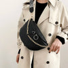A woman wears a beige trench coat and carries a black woven pattern waist bag with a gold chain strap.