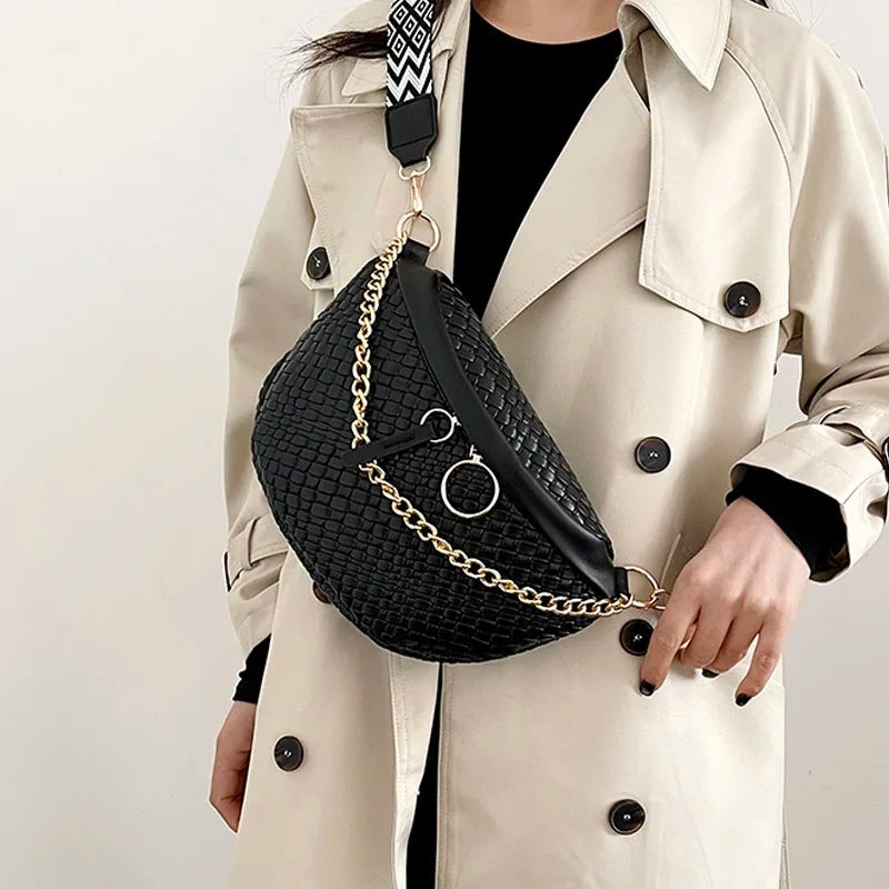 A woman wears a beige trench coat and carries a black woven pattern waist bag with a gold chain strap.