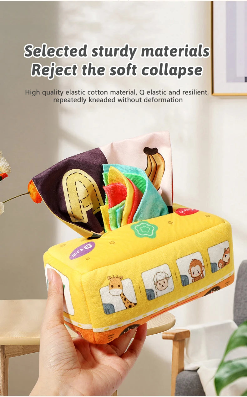 A hand holds a yellow sensory tissue box toy with colorful crinkle scarves and fabric pieces.