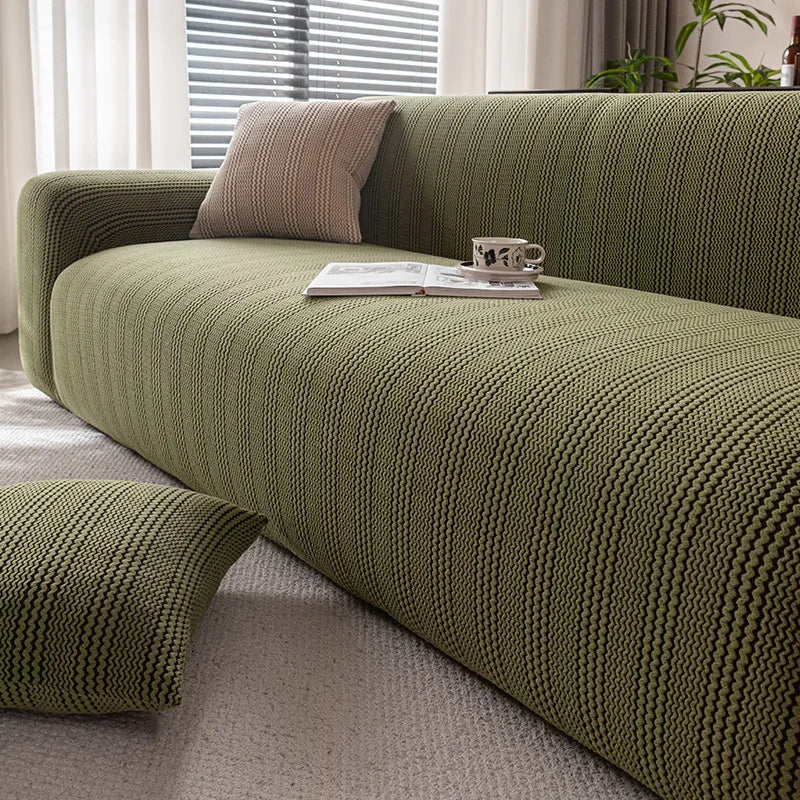 A textured olive green quilted stretch sofa cover with a neutral throw pillow and an open book on the surface.