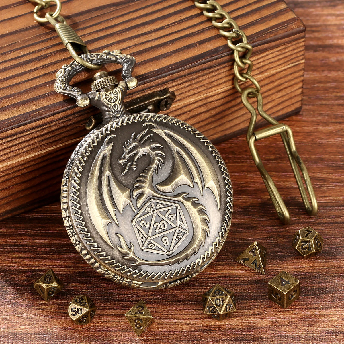A brass dragon-themed pocket watch case with a chain and seven metal polyhedral dice resting on a wooden surface.