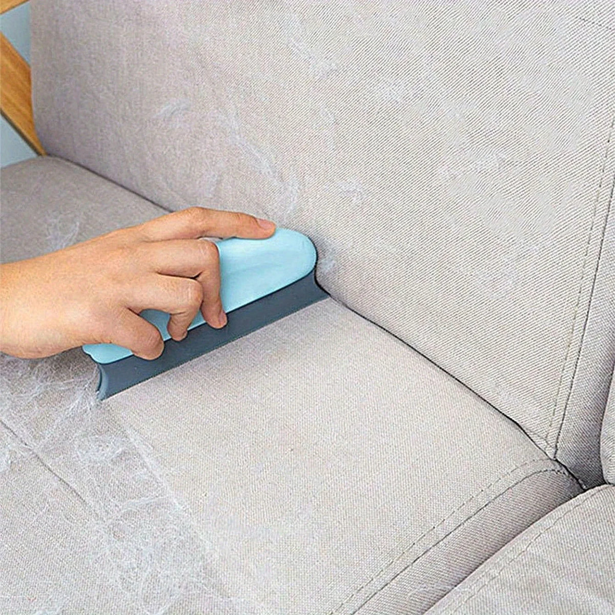 A hand is using a blue pet hair removal brush on a gray sofa fabric.