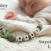 A green and beige beaded pacifier clip necklace with a heart charm rests on a white blanket next to a baby's hand.