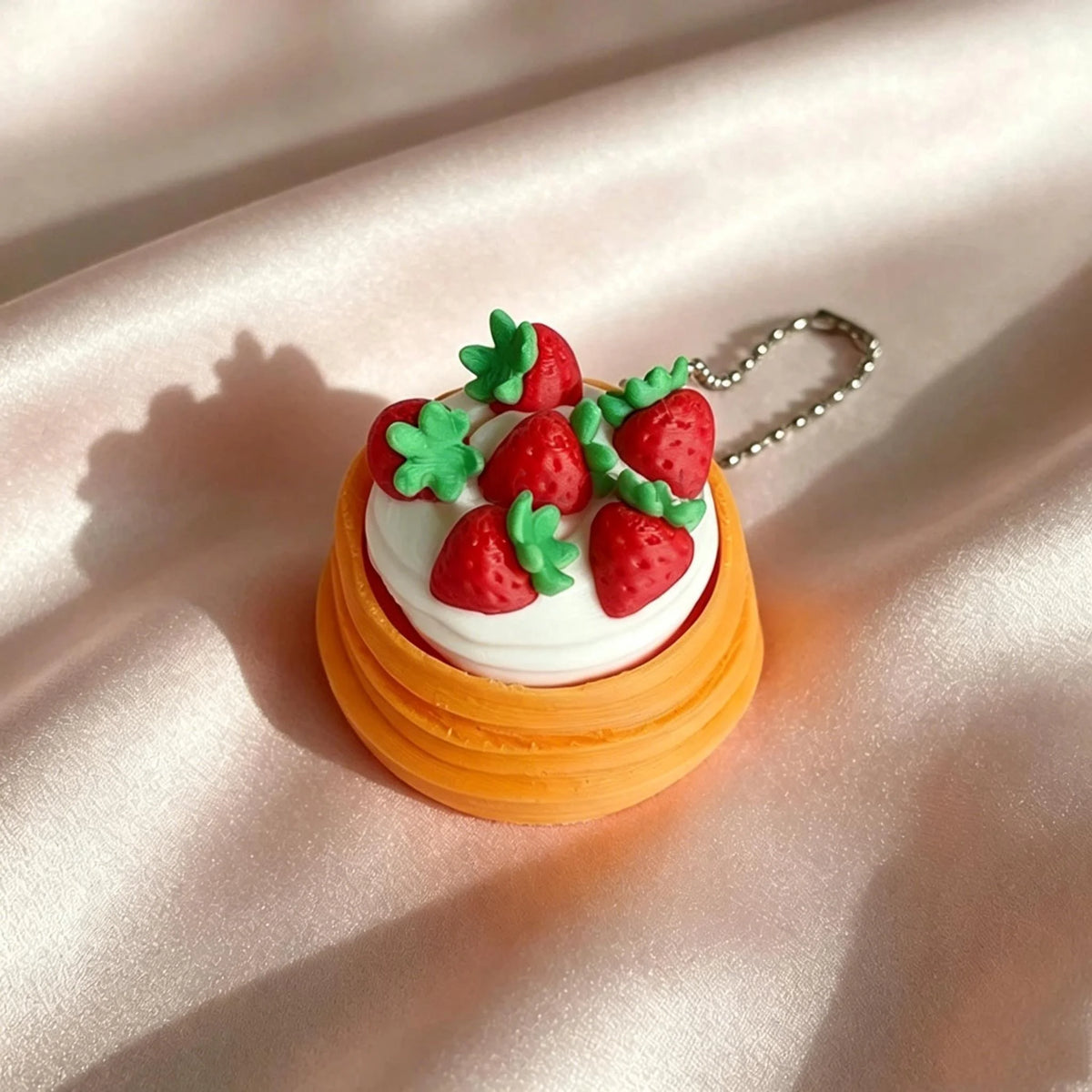 A small, round cake decorated with white frosting and fresh strawberries is attached to a silver keychain.