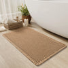 A rectangular beige waffle weave bath mat with tassels placed on a light wood floor next to a white bathtub.
