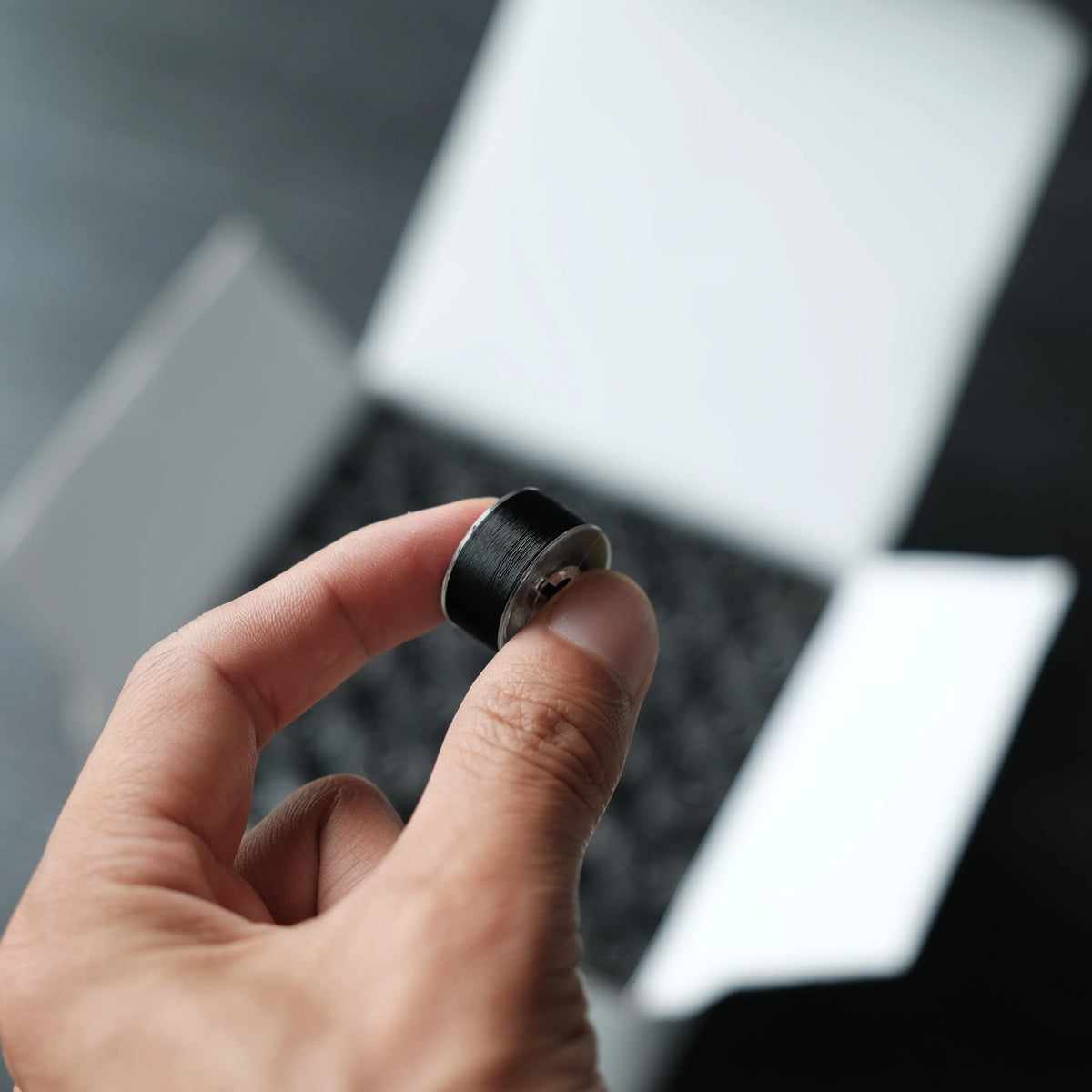 A hand holds a black bobbin with a silver thread for home embroidery machines.