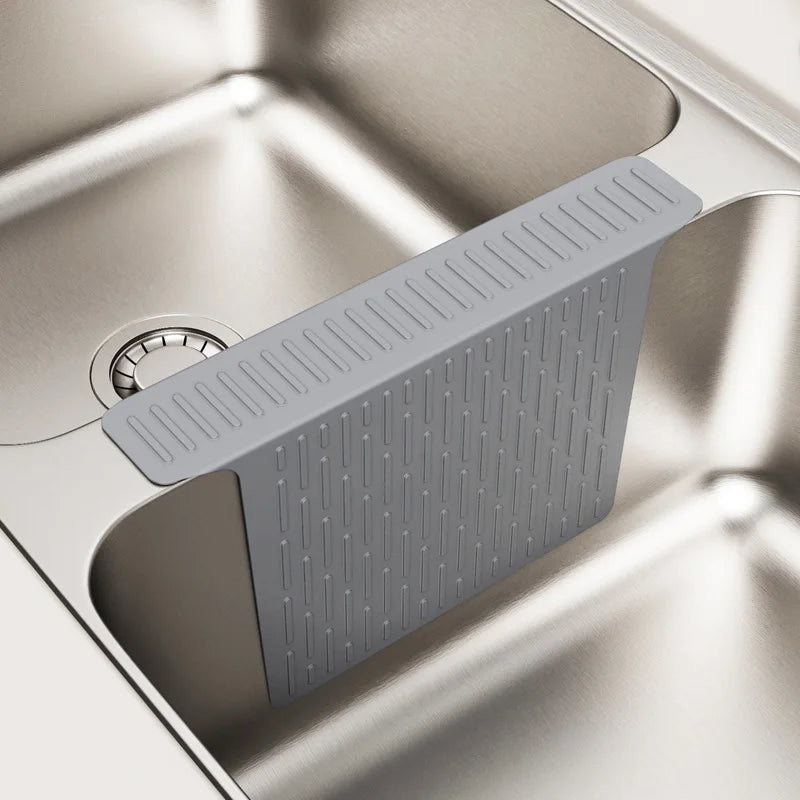 A silicone sink protector mat with drainage holes is shown in a close-up view inside a stainless steel kitchen sink.