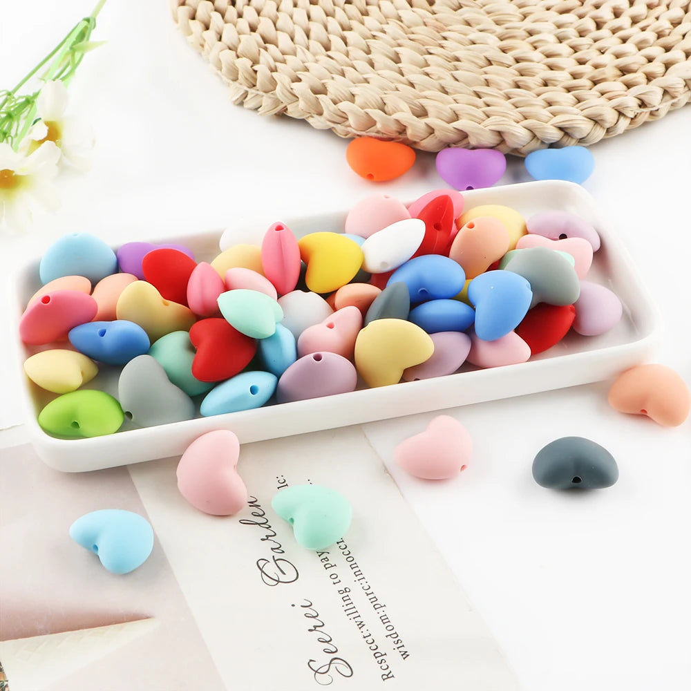 A collection of ten colorful silicone heart beads in various shades are displayed on a white tray.