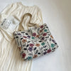 A beige corduroy tote bag with floral accents resting on a white dress and clear glass.