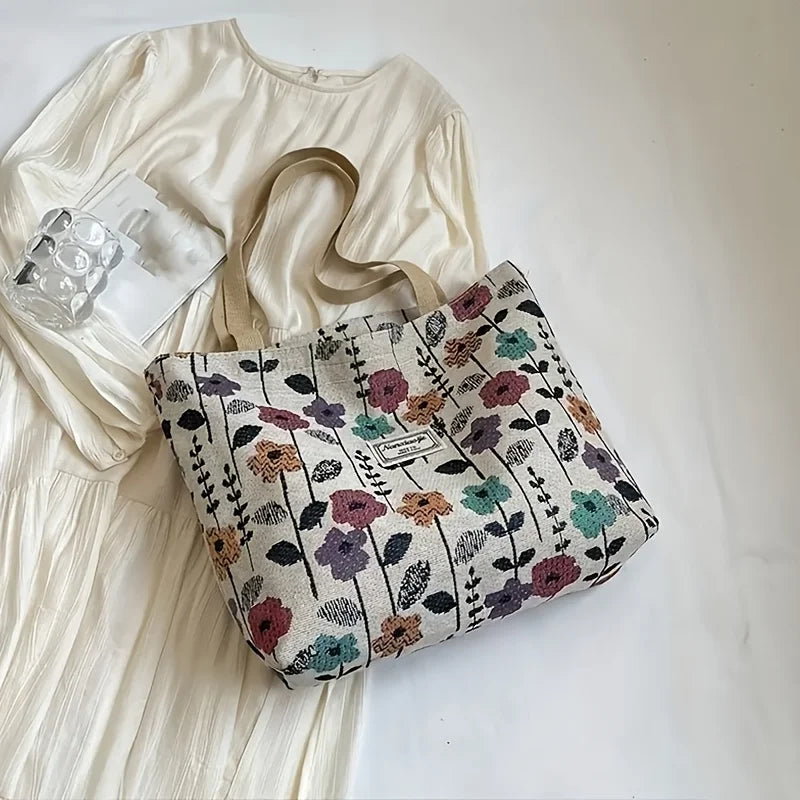 A beige corduroy tote bag with floral accents resting on a white dress and clear glass.