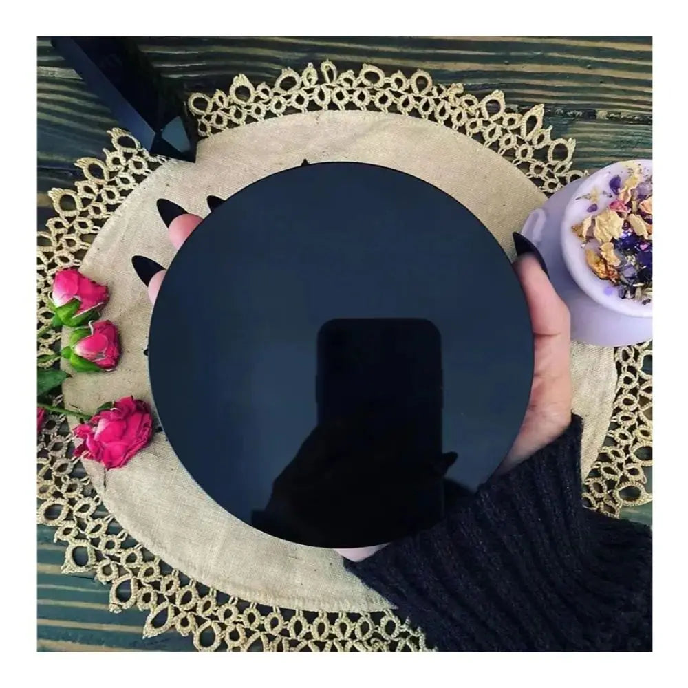 Une main tient un miroir de divination en obsidienne noire, posé sur un tapis tissé avec des bordures de dentelle.