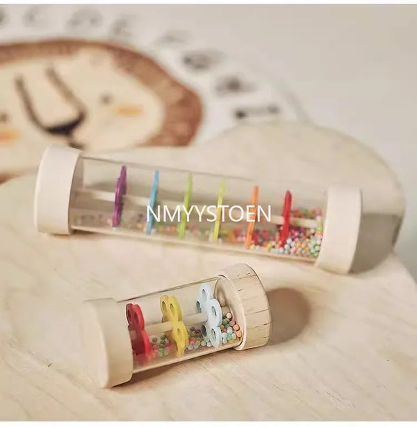 A clear wooden rain sound pipe toy filled with colorful beads resting on a light wooden surface.