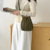 A woman wearing a cream sweater and white skirt holds a olive green pleated faux leather crossbody bag while using a laptop.