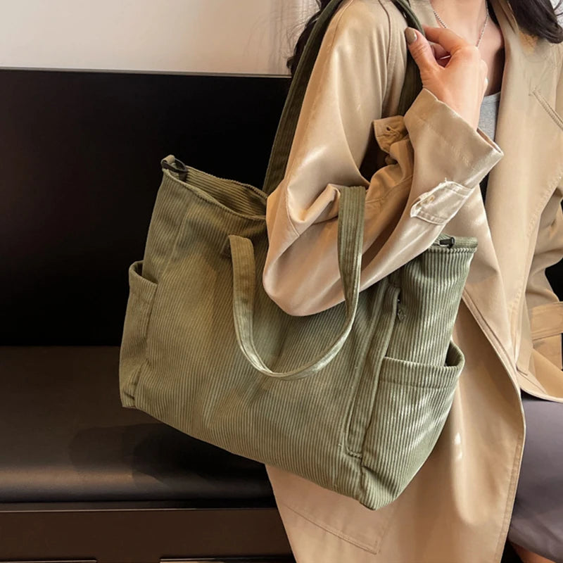 A woman holds a light beige jacket and a green corduroy tote bag with an adjustable strap.