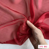 A hand is folding a piece of sheer, reddish-pink silk chiffon fabric.