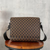 A brown patterned crossbody bag resting on a wooden stool against a gray concrete background.