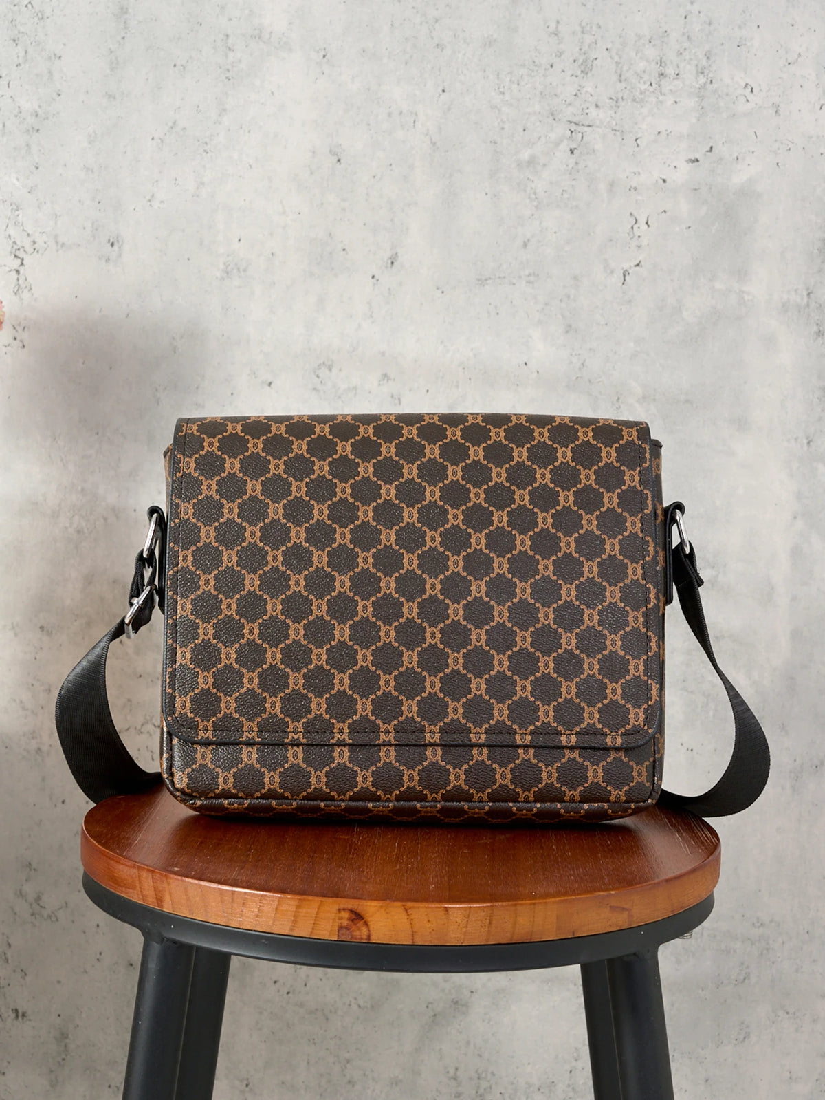 A brown patterned crossbody bag resting on a wooden stool against a gray concrete background.