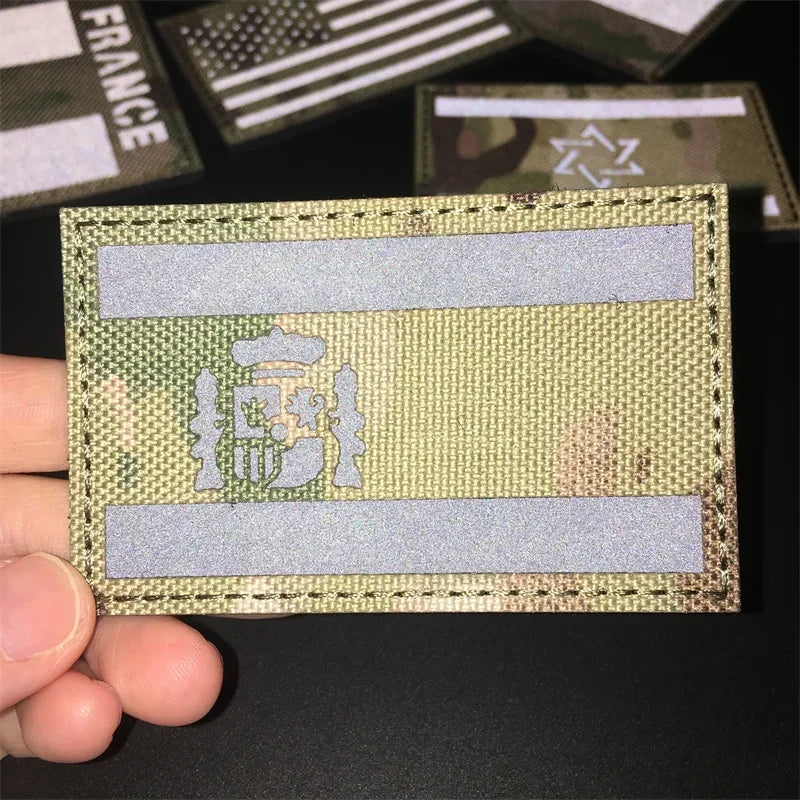 A hand holds a rectangular tactical patch featuring a national flag design with hook-and-loop backing.