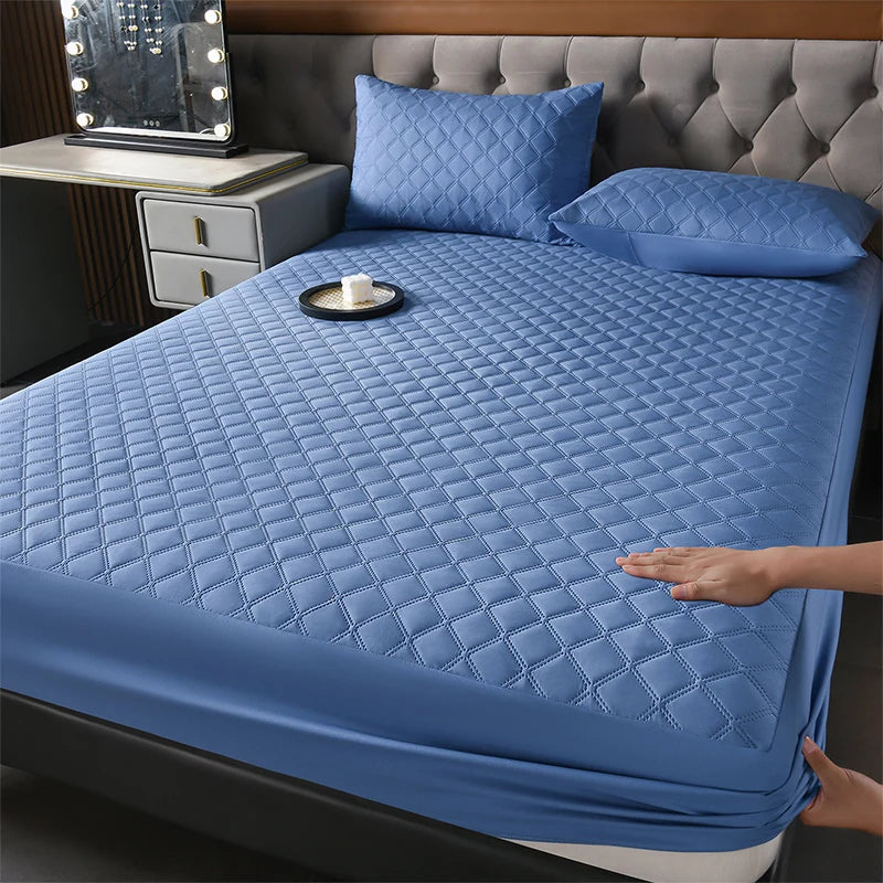 A blue quilted waterproof mattress protector is shown on a bed, with matching pillows and a bedside lamp visible.