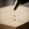 A drop of clear glue is being dispensed from a hot melt glue gun onto a wooden surface.