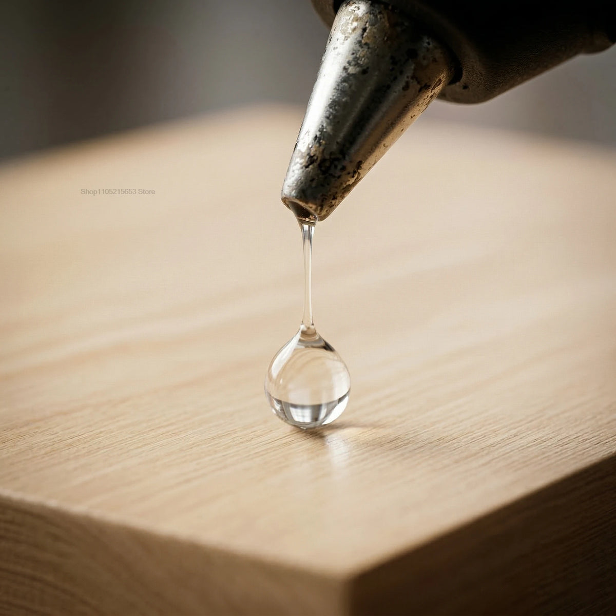 A drop of clear glue is being dispensed from a hot melt glue gun onto a wooden surface.