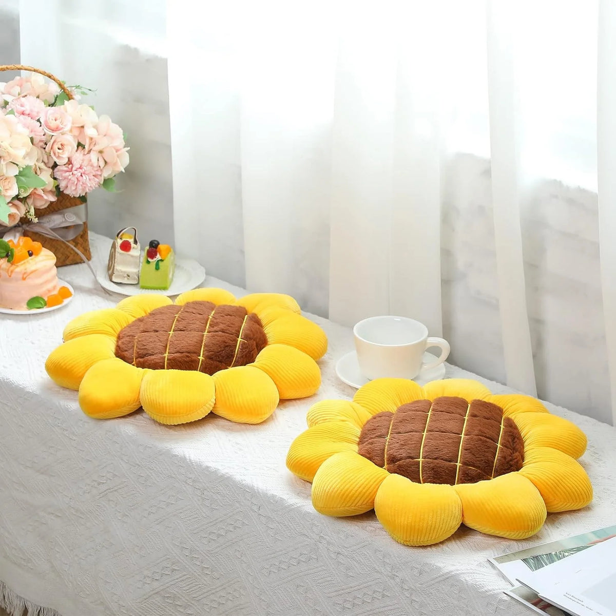 A collection of yellow sunflower-shaped pillows arranged on a white cloth with pink flowers and a cup in the background.