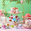 A collection of Sylvanian Families tea party baby figures in various outfits and accessories displayed on a gold stand.
