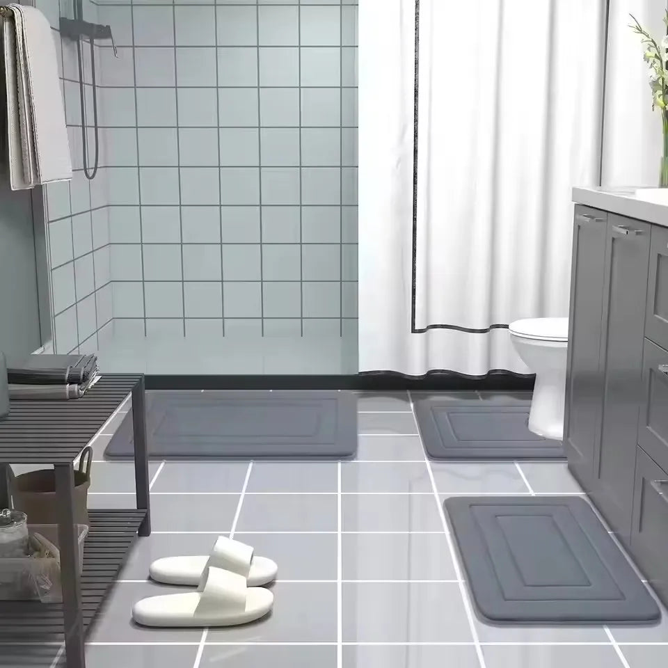 A collection of gray non-slip bathroom mats and white memory foam slippers are displayed on a tiled floor.