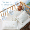 White ice silk baby pillow with a brown teddy bear toy shown in a wooden crib setting.