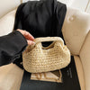 A woven straw chain crossbody handbag resting on a black surface next to a person's hand.