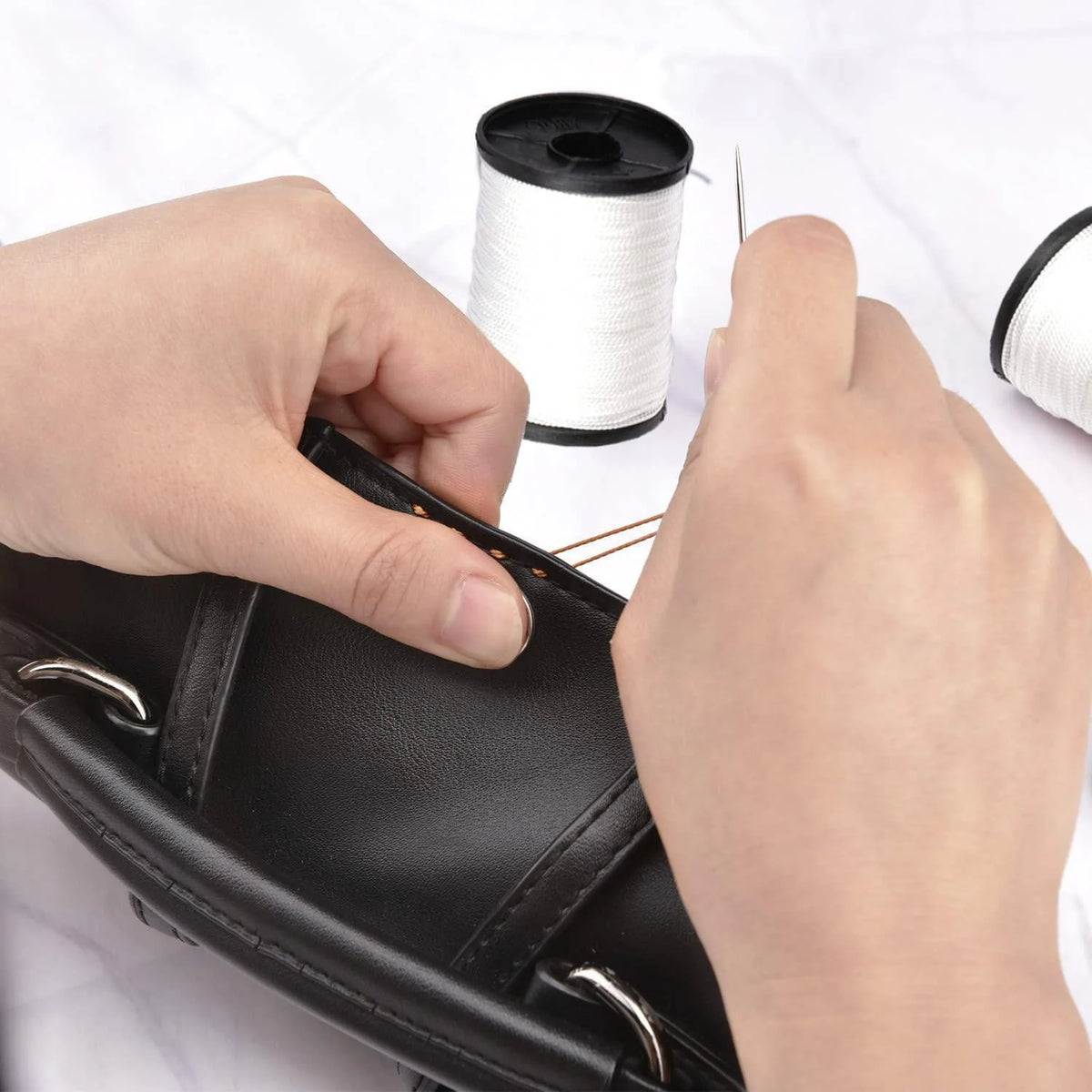 Hands are using a sewing needle and heavy duty nylon thread to repair black leather upholstery.