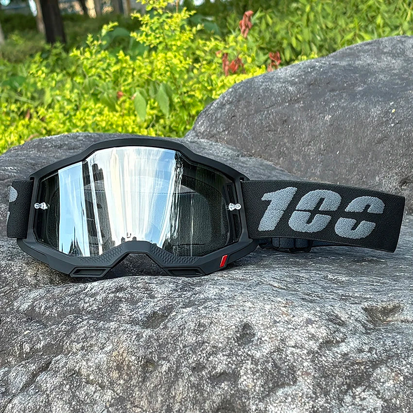 A pair of black motocross goggles with a gray strap resting on a large gray rock.
