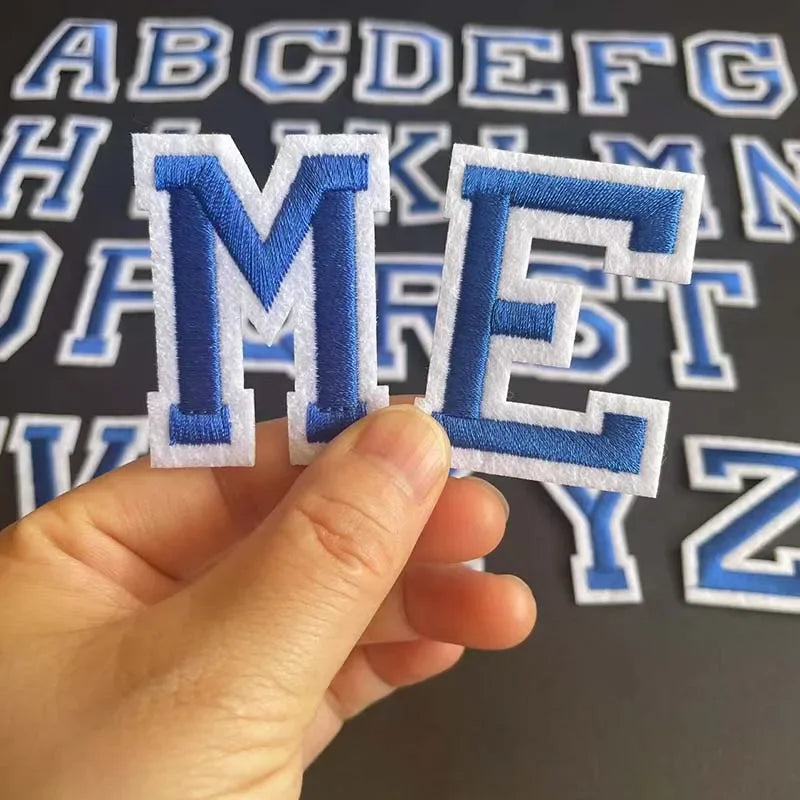 A hand holds an embroidered blue and white alphabet patch letter M and E from a set.