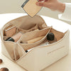 A hand is opening a beige leather travel makeup organizer bag, revealing various cosmetic items inside.