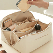 A hand is opening a beige leather travel makeup organizer bag, revealing various cosmetic items inside.