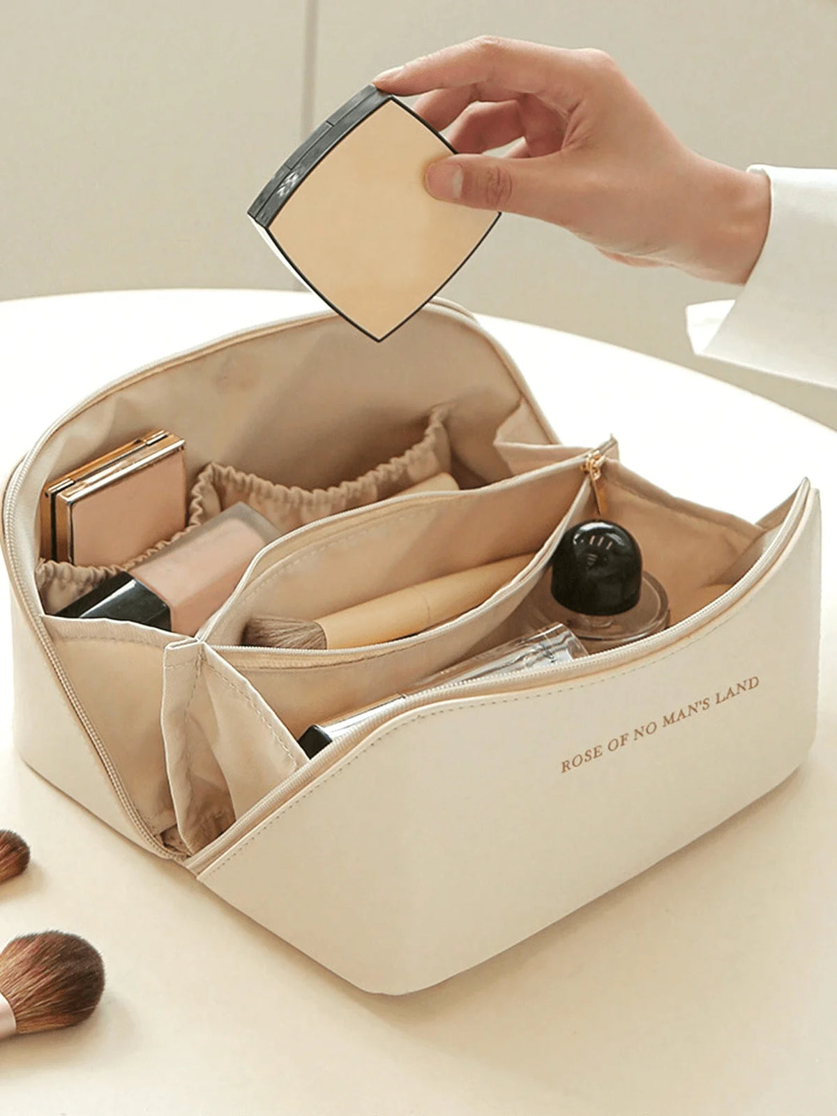 A hand is opening a beige leather travel makeup organizer bag, revealing various cosmetic items inside.