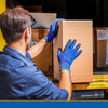 A man wearing blue work gloves is handling a cardboard box with a light brown background.