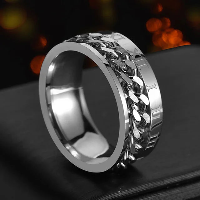 A close-up shot of a stainless steel spinner ring with a textured, chain-link design resting on a dark surface.