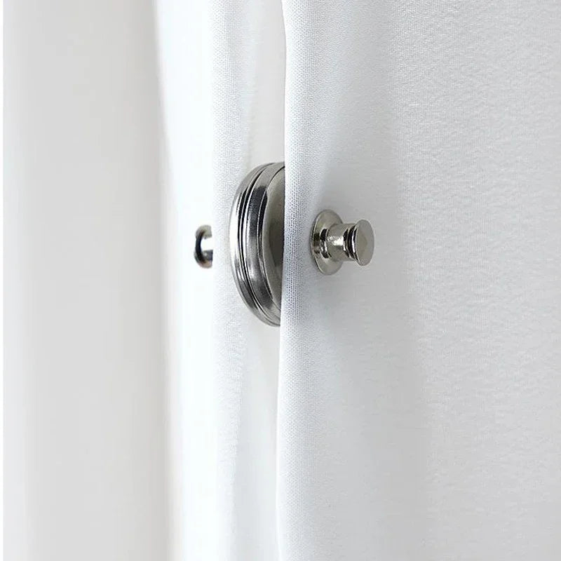 Close-up of a white curtain with a silver magnetic clip and button fastener on the fabric.