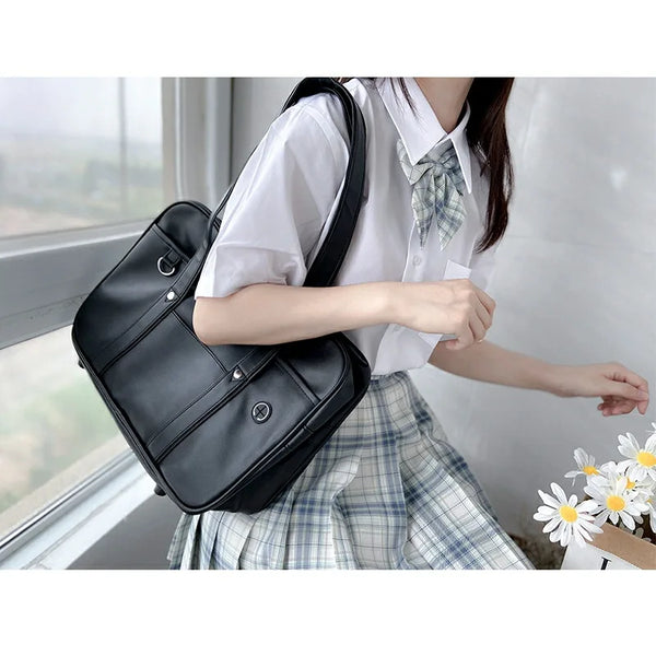 A woman wearing a white shirt and plaid skirt holds a black PU leather shoulder bag by a window.