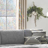 A gray sofa in a bright room with a window featuring rainbow stained glass and a green leafy hanging decoration.