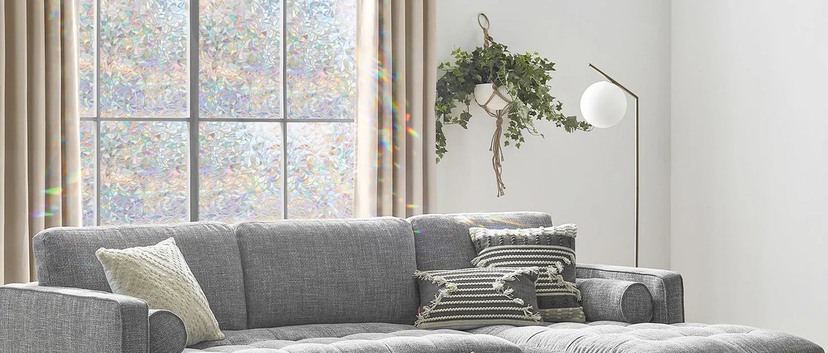 A gray sofa in a bright room with a window featuring rainbow stained glass and a green leafy hanging decoration.