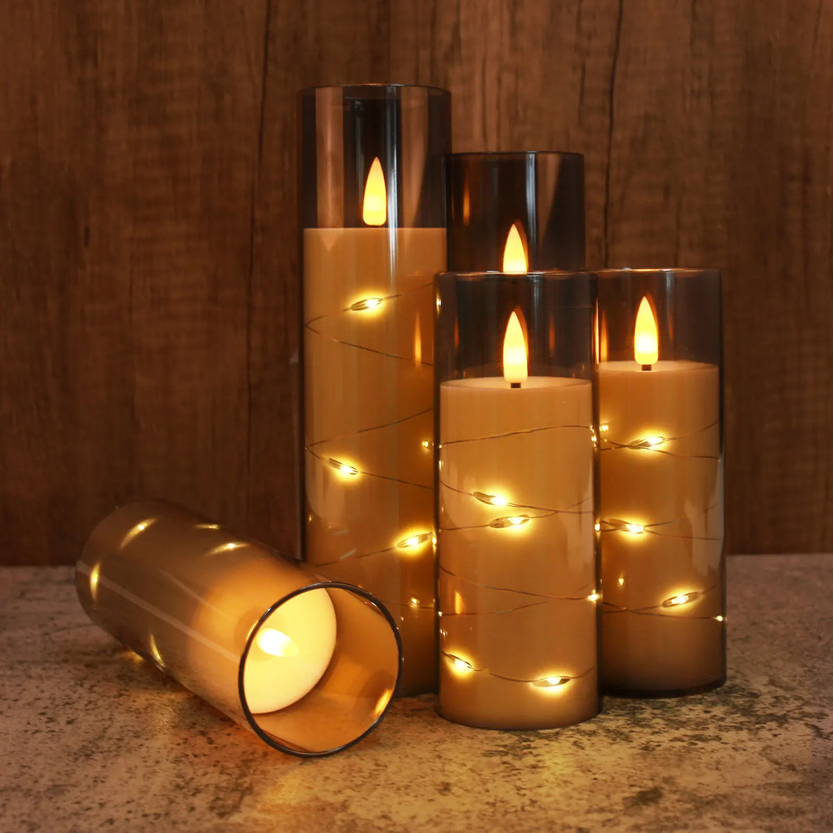A set of three tall glass candles with flickering LED flames and twinkling string lights on a stone surface.