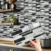 A hand holds a rectangular tile sticker for kitchen backsplash against a dark gray and white marble subway tile wall.