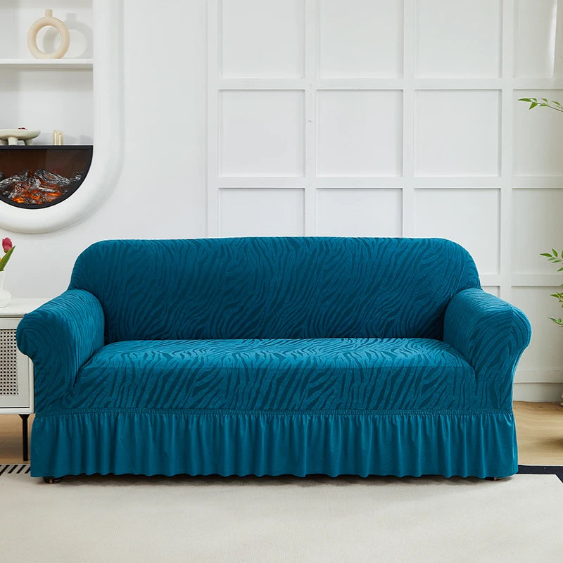 A teal jacquard sofa cover with an elastic skirt displayed on a white couch against a paneled wall.