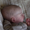 A close-up of a realistic 19-inch silicone vinyl reborn baby doll with soft skin and light brown hair, resting in a textured 