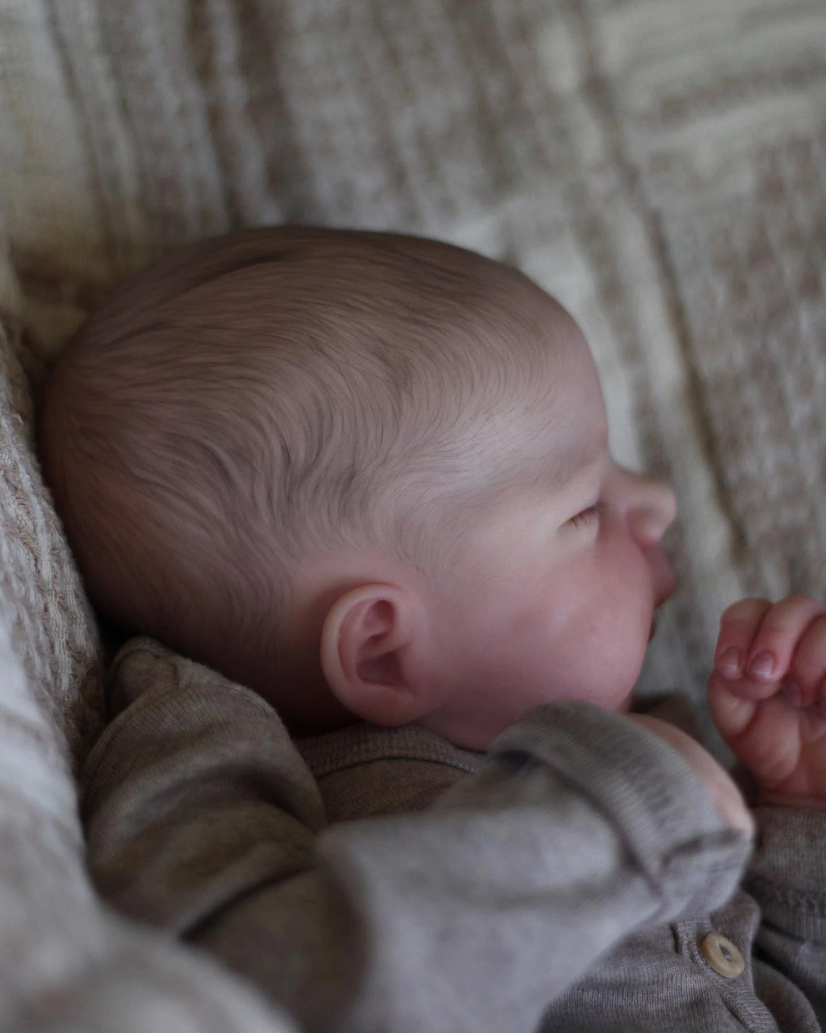 A close-up of a realistic 19-inch silicone vinyl reborn baby doll with soft skin and light brown hair, resting in a textured 